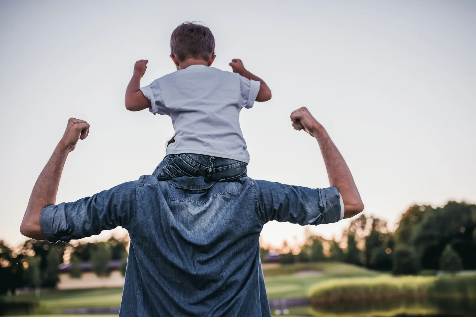 Father carrying his young child on his shoulders outdoors, both with arms raised